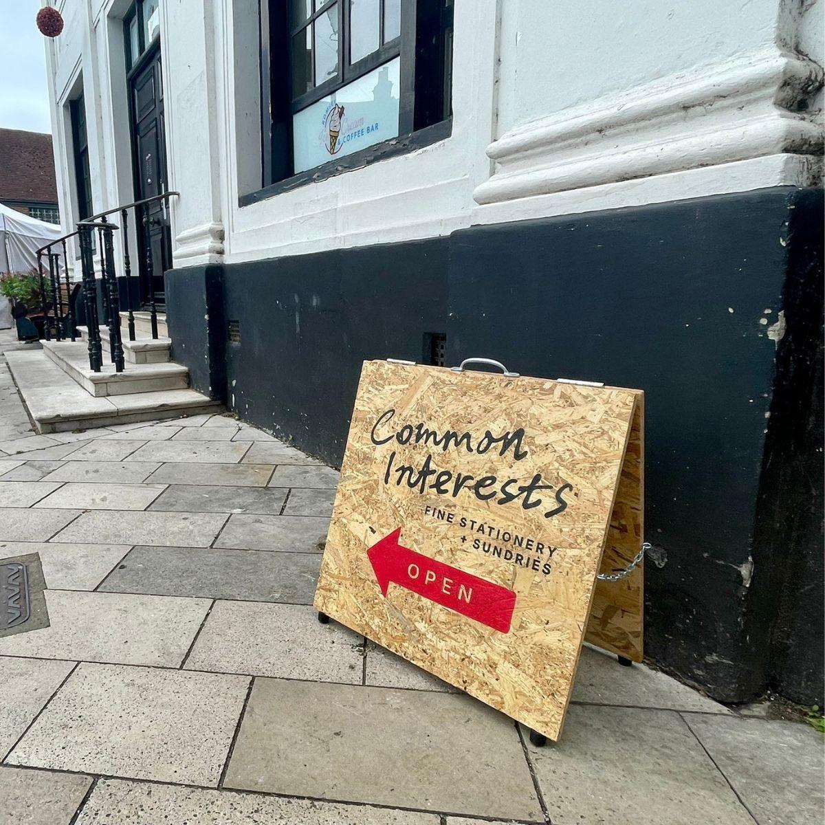 A custom wooden A-frame sidewalk sign for Common Interests Fine Stationery + Sundries, featuring a red arrow marked OPEN on a stone-paved street next to a storefront.