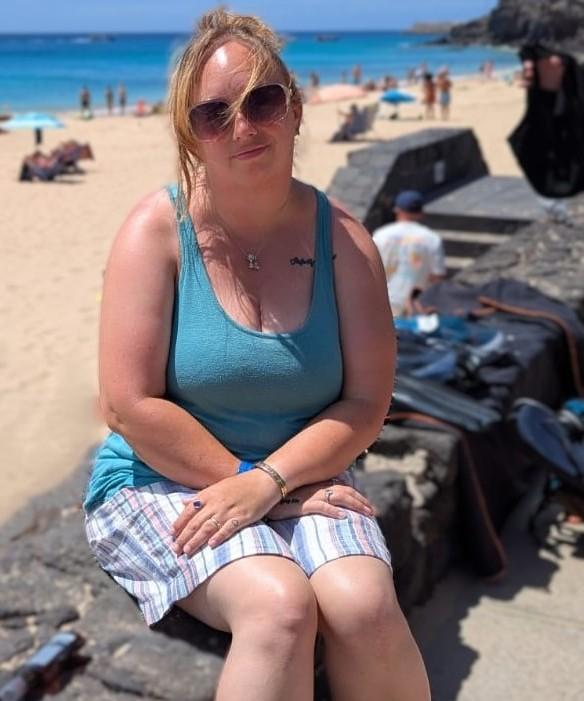 Lanie, founder of Drops of Wild Raine, sitting on a stone wall at a sunny beach wearing sunglasses and a turquoise top. A calm ocean and sandy beach stretch out behind her
