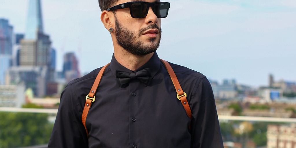 A brown leather harness with gold hardware worn by a male model on top of a black shirt at outdoor location in London with The Shard Tower in the background.