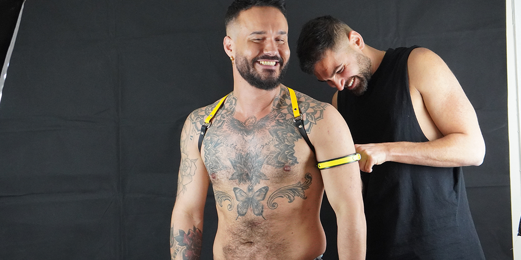 A behind-the-scenes image showing a shoulder harness and arm band in Black and Yellow leather, worn by a male model with a stylist in the back helping the model. Both are laughing. 