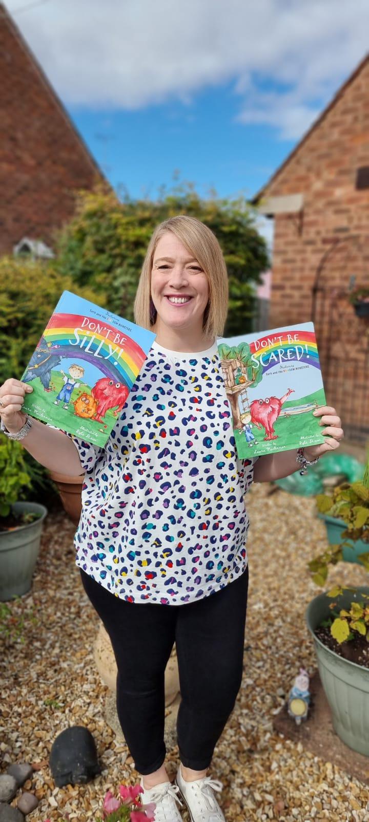Lozza Mac stood outside in a rainbow leopard print t-shirt holding her first two books, Don't Be Silly! and Don't Be Scared!