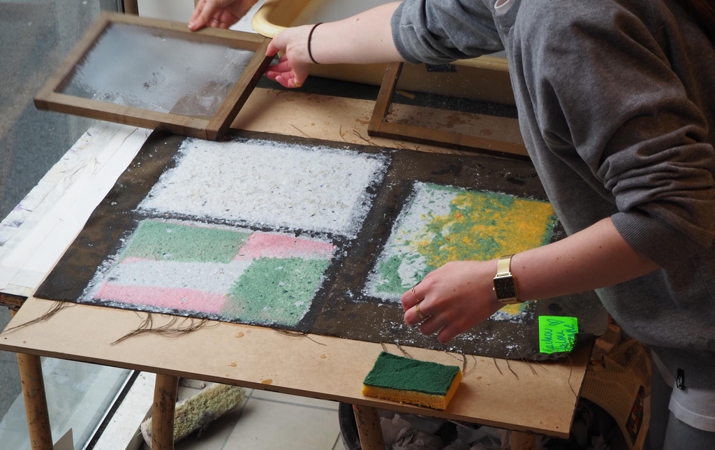 Mise en scène de la fabrication du papier recyclé avec la forme à papier et des feuilles couchées sur un feutre étendue sur une table.