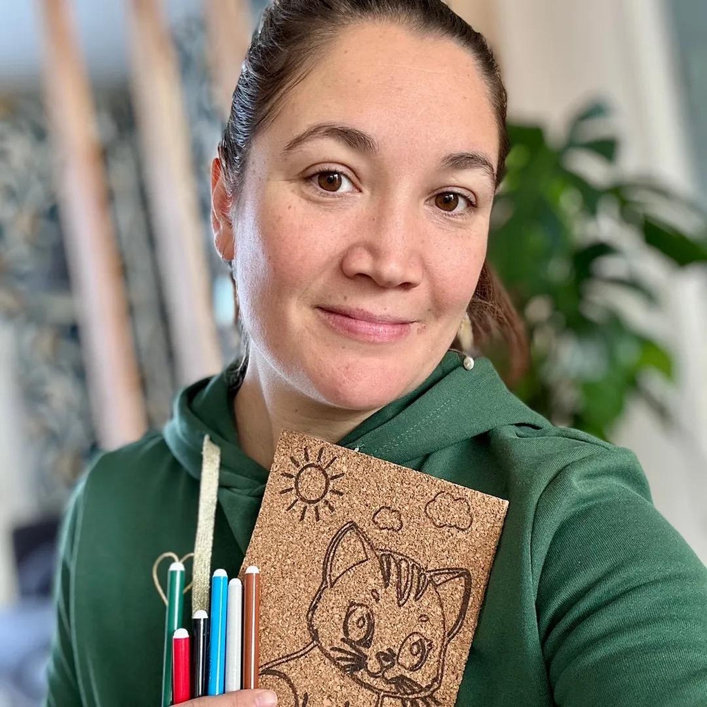 Hélène créatrice de Rebelote. Elle sourit, porte un sweet à capuche vert et tient dans sa main une plaque à colorier Rebelote et des feutres