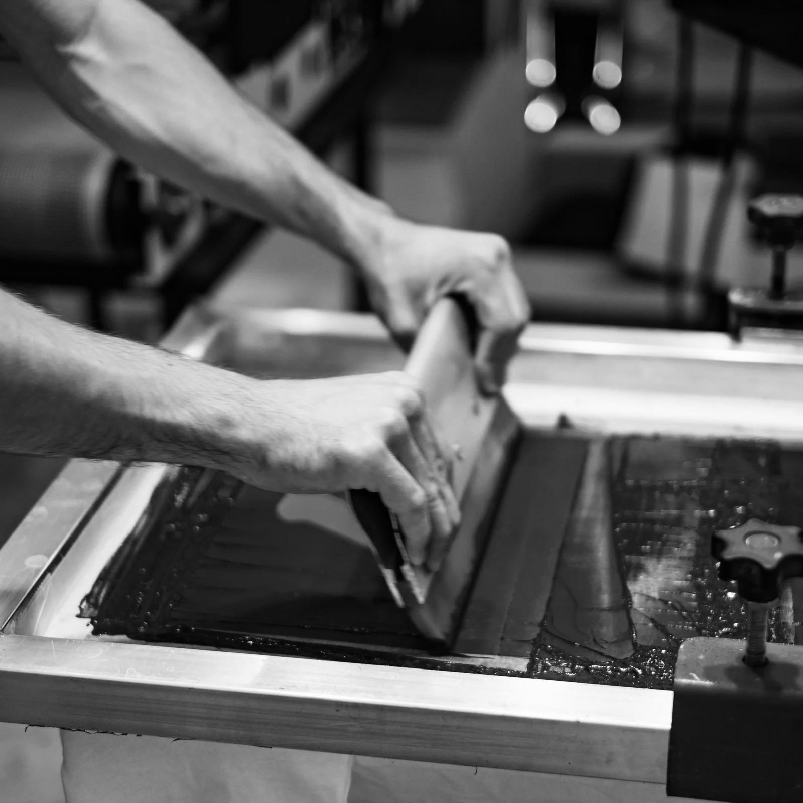 Gros plan en noir et blanc sur des mains en pleine sérigraphie artisanale, une technique qui incarne l'essence du savoir-faire de Vestimentari. Chaque t-shirt, pull et casquette de la marque est sérigraphié à la main à Arles, soulignant un engagement fort pour l'artisanat local et la qualité. Vestimentari s'inspire des traditions provençales pour créer des vêtements intemporels, minimalistes et fonctionnels, pensés pour le quotidien. Cette image met en lumière le processus créatif et la passion derrière chaque pièce, reflétant l'authenticité et l'élégance de la Provence moderne.