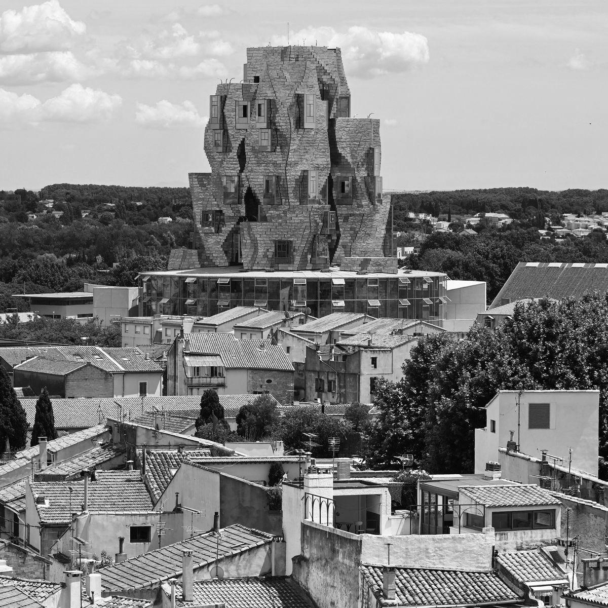 Photo en noir et blanc de la tour emblématique LUMA à Arles, surplombant les toits typiques de la ville. Cette fusion architecturale entre tradition et modernité reflète parfaitement l'esprit de Vestimentari, une marque inspirée par la Provence. Tout comme la tour LUMA réinvente le paysage d'Arles, Vestimentari réinvente les vêtements du quotidien avec des t-shirts, pulls et casquettes intemporels et minimalistes. Fabriqués avec soin, ces produits incarnent l'élégance fonctionnelle et l'héritage provençal, mêlant savoir-faire local et design contemporain. Une célébration de l'authenticité et de la modernité.