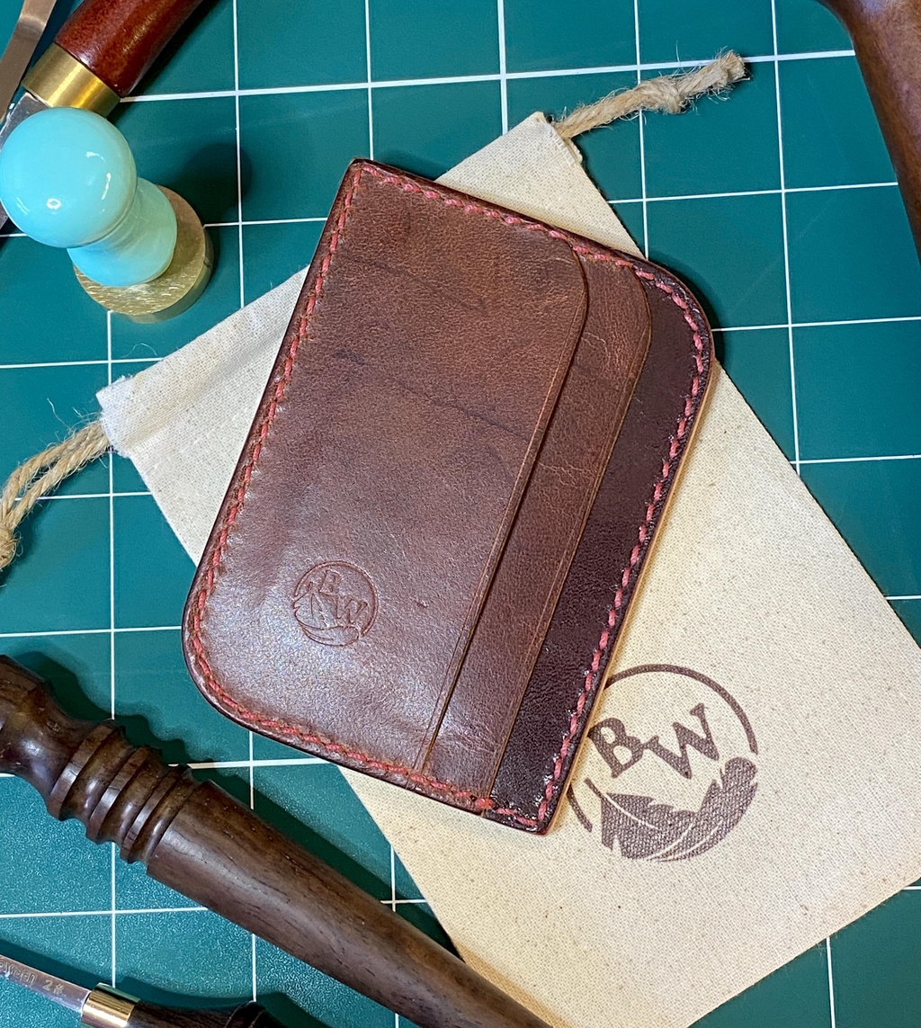 Background photo of a Burgundy and Copper Beech coloured 'Haymarket' card holder with red stitching, that is laying on a natural linen dust bag with the Broken Wing Feather logo on a workbench, next to leathercraft tools.

Headline reads: Natural Materials, Traditional Methods

Text reads: "Every order is individually cut, assembled, saddle-stitched, and finished by hand."

There is a button marked 'Explore' that can be clicked on to access the product range.