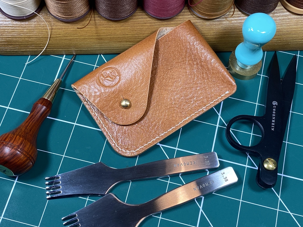 A photo showing a tan coloured calfskin 'Dalry' card holder on a green cutting mat, surrounded by artisan leatherwork tools, and waxed thread.

Headline reads: "Come with us, on a journey back to the 'old school'..."

Text reads: "Here at Broken Wing Leathercraft Studio we have a serious passion for making things with our hands, and doing it all the old-fashioned way. And while using these natural materials and traditional methods may take longer (and be much more expensive for us to produce!), we just can't get away from how it feels to slow down and do things 'properly' in a world that is constantly demanding quick results and instant gratification. 

For us, the patina of well-worn leather, its natural aroma, the lovely sound a Sam Browne button makes against leather when you fasten and unfasten it, and the tactile joy of full-grain veg-tan leather, is all so deeply satisfying to the senses. So, if you want a humble return those elusive slow moments, join us, and enjoy your very own piece of natural craftsmanship to enrich the  mundanity of everyday life!"