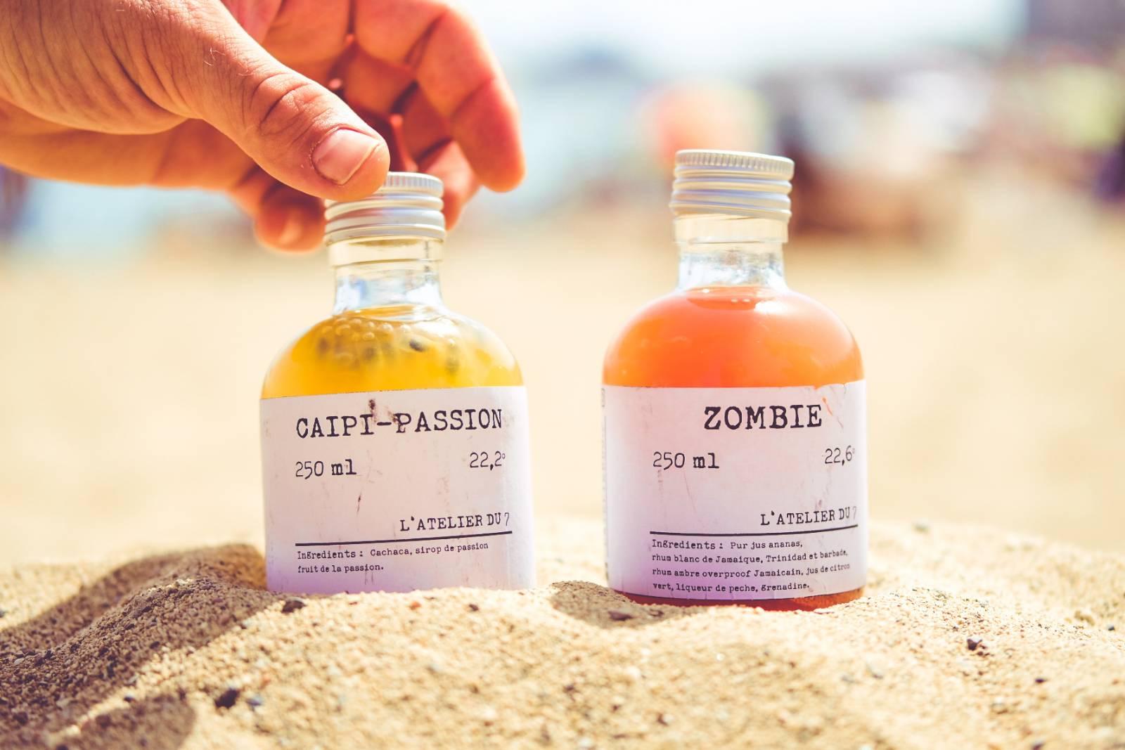Deux mini-bouteilles de cocktails L'Atelier du 7, Caïpi-Passion et Zombie, posées sur du sable