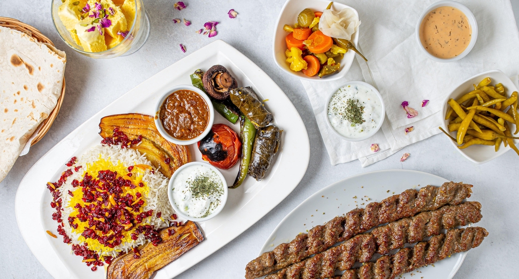 Mehrere Teller mit drei Lammhackspießen, gegrillte Auberginen, Tomaten und Peperonis und Reis auf weißem Hintergrund.
