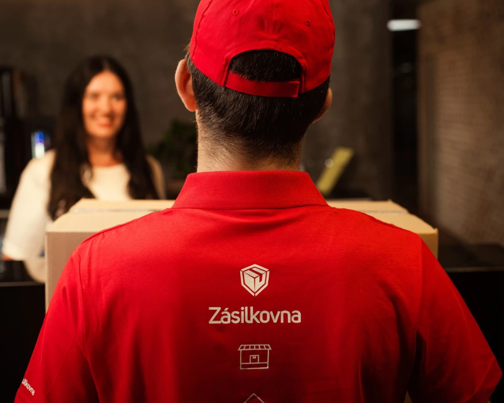 Na fotografii je kurýr Zásilkovny v červeném tričku s červenou kšiltovkou předávající balíčky ženě s černými vlasy.