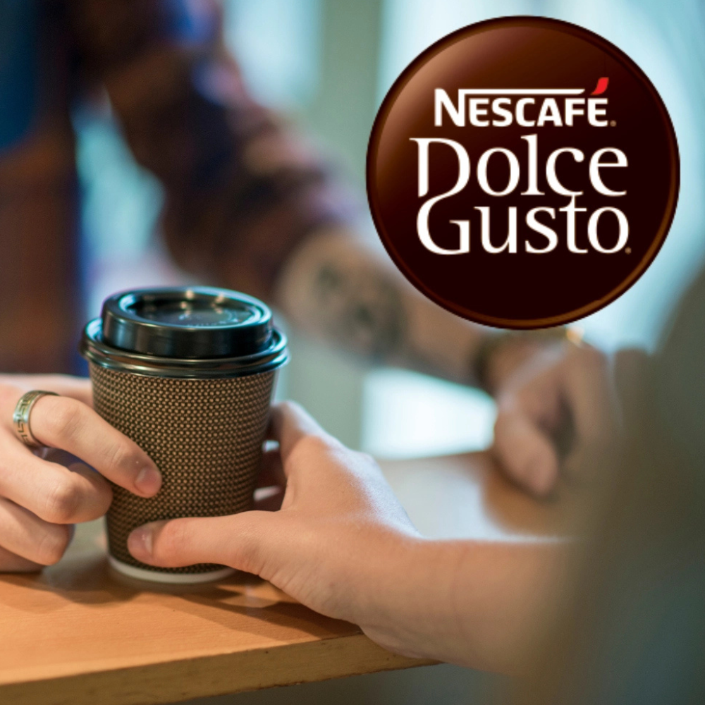 Na fotografii je ženská ruka předávající kelímek na coffeeto go pánské ruce, pře hnědý pult.