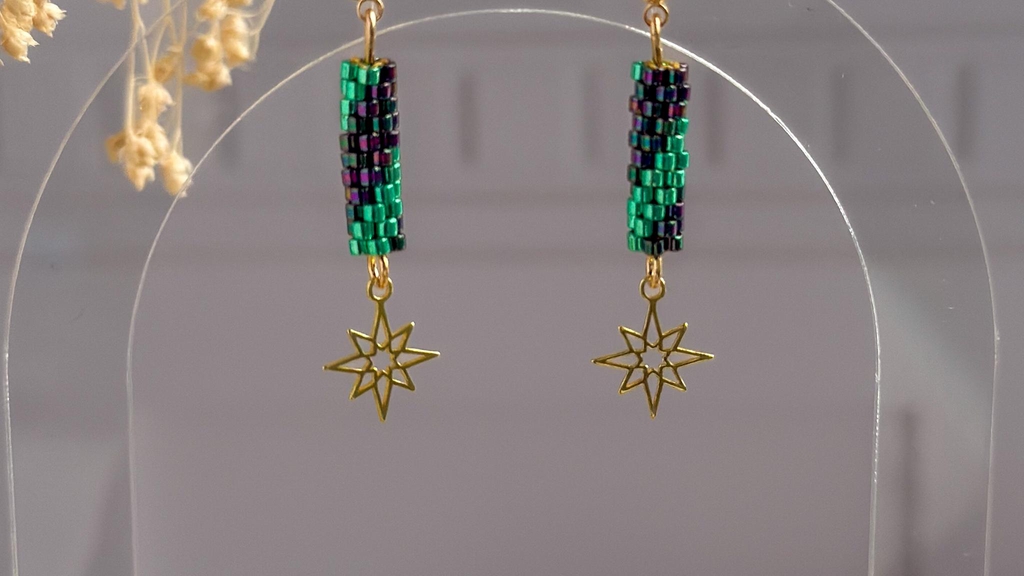 Boucles d'oreilles tissées main Élycea avec perles Miyuki irisées vert et violet et breloque étoile dorée, suspendues sur fond gris avec fleurs séchées