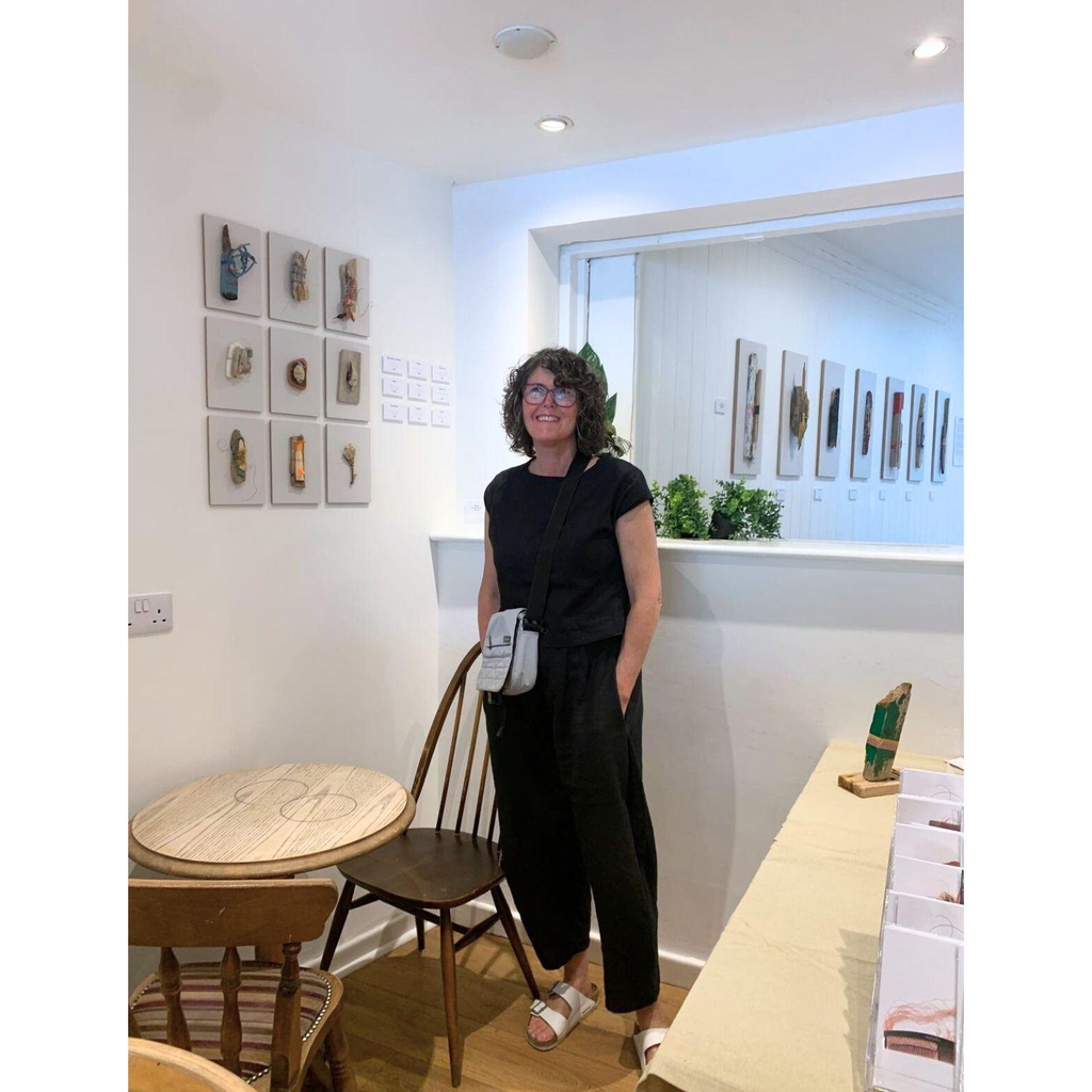 3D eco freindly wall art | assemblage and sculpture | The image shows the Leeds based artist, Jane Wood standing in a room with a display of 9 small pieces of wall art on the wall to the left with seven larger artworks on the wall behind her.  The exhibition is called Reshaping The Past.