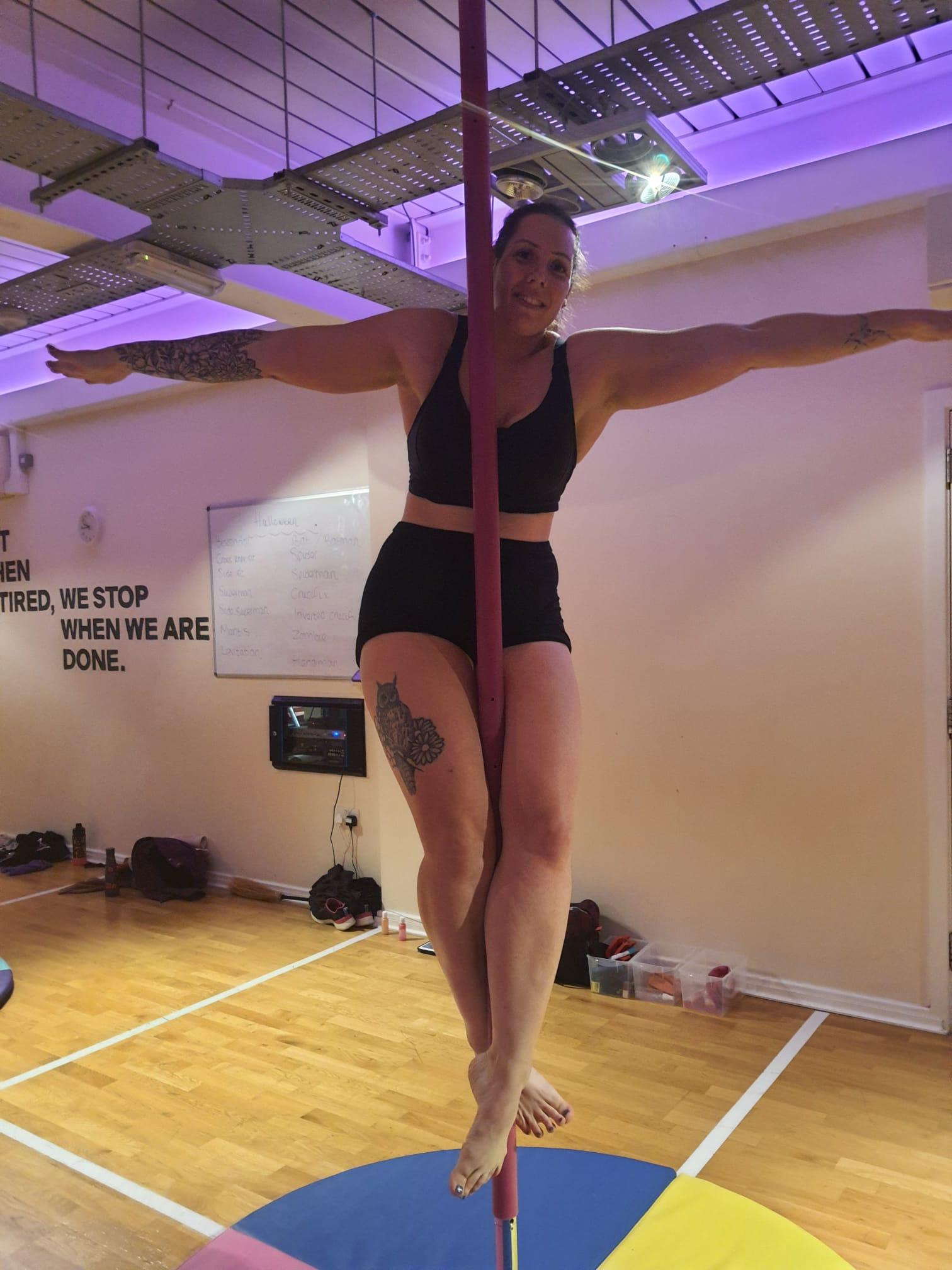 Woman wearing black shorts and sports bra, performing a standing crucifix pole fitness move on a pink silicone pole