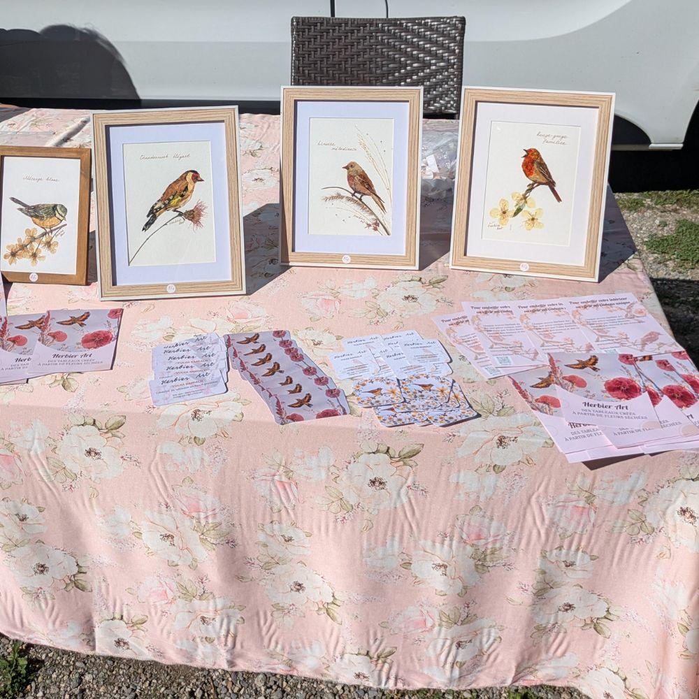 Stand Herbier Art à la Fête des Bûcherons au Fayet, avec quatre tableaux encadrés d’oiseaux créés en fleurs pressées, des cartes et des brochures.
