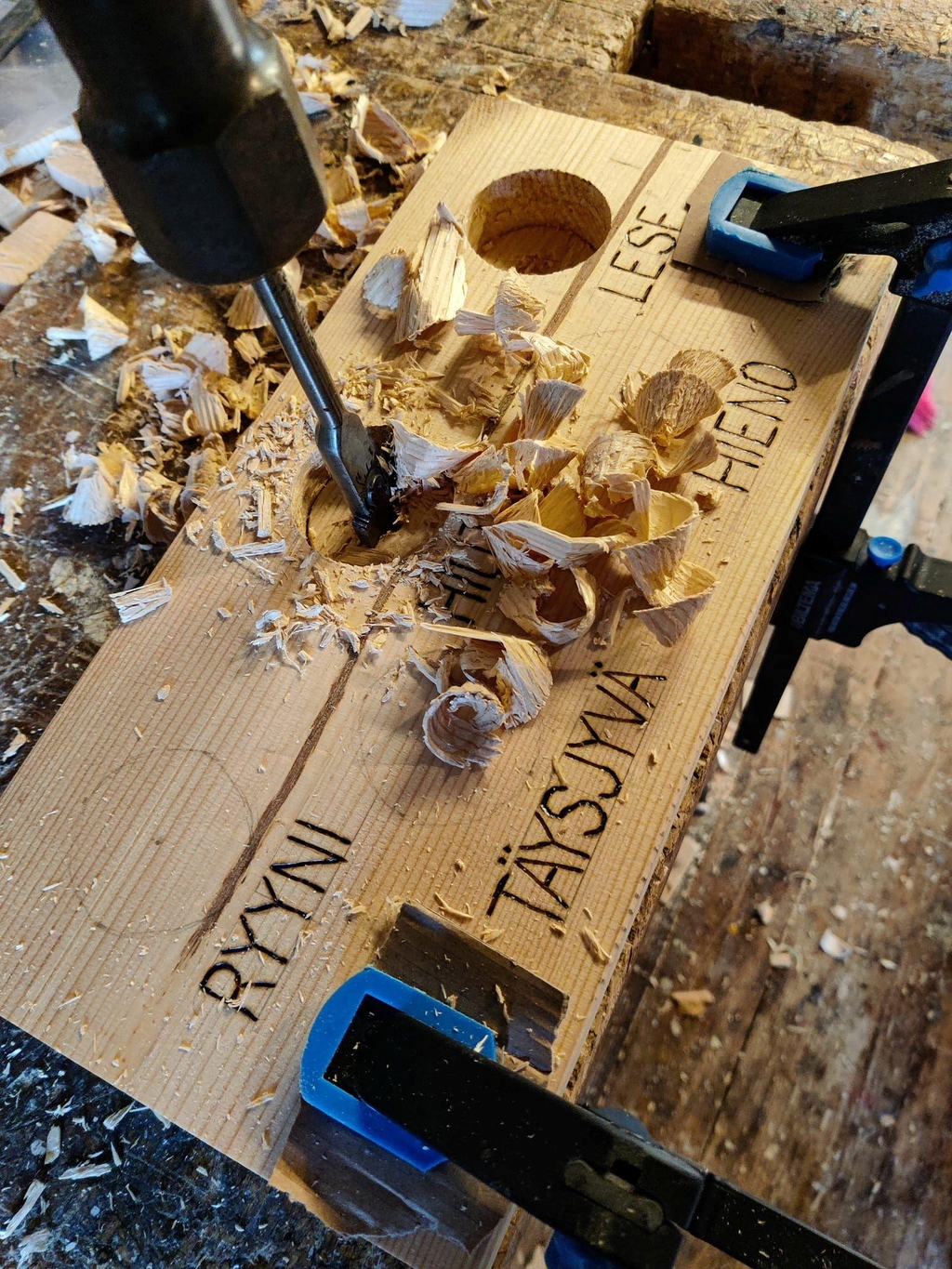 Käsityönä työstetty tuotekyltti, jossa on tekstiä, puulastuja ja käsityökaluja.