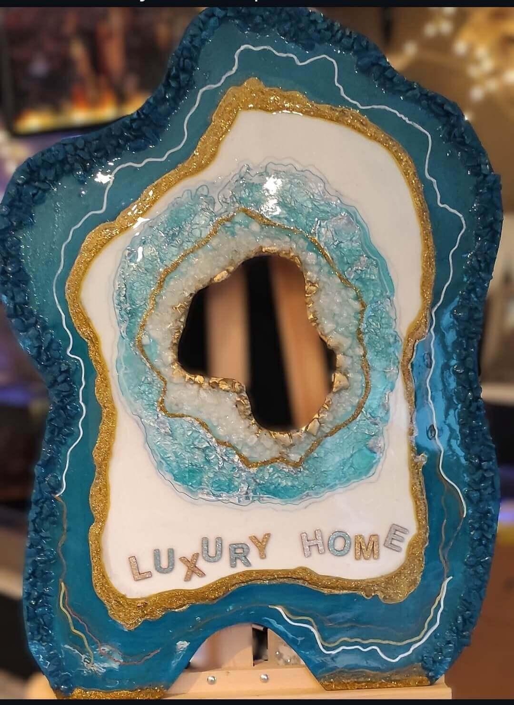 Tableau à poser. Création artisanale en forme de géode d'agate dans les tons bleu canard, blanc et doré, avec l'inscription "LUXURY HOME". La pièce présente un centre évidé texturé de cristaux et des lignes dorées scintillantes.Décoration artistique faite main inspirée d'une géode naturelle. L'objet aux contours irréguliers arbore des nuances de bleu océan, de blanc et de feuilles d'or.
