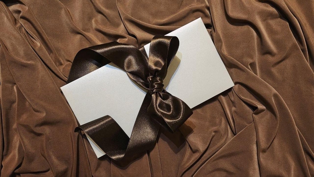 carte cadeau couleur ivoire entouré d'un jolie ruban satiné marron en forme de noeud. Le tout étalé sur un tissu brillant marron.