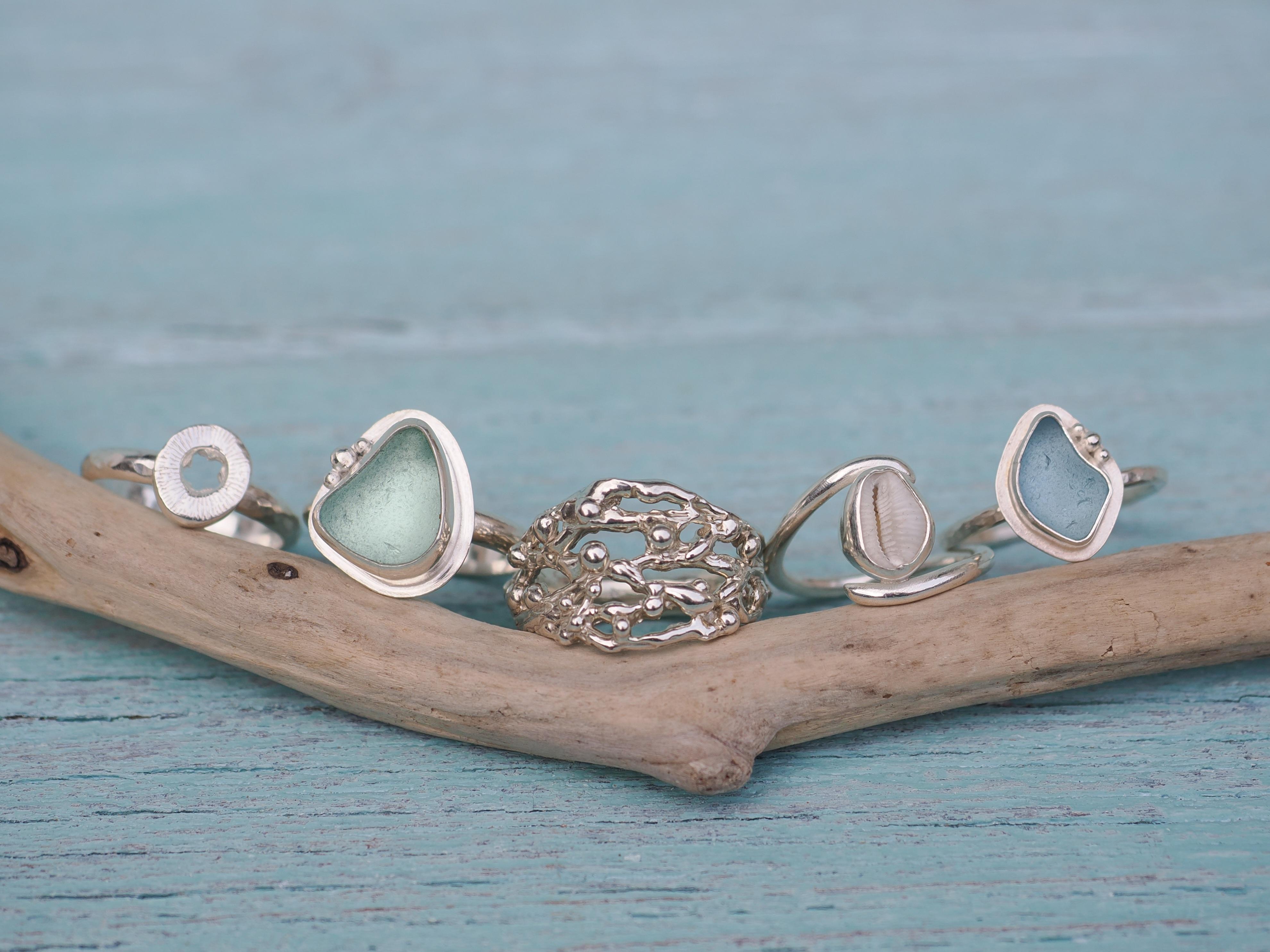 Rings resting on driftwood: sea glass, cowrie, silver Seaweed and a silver cuddy bead ring