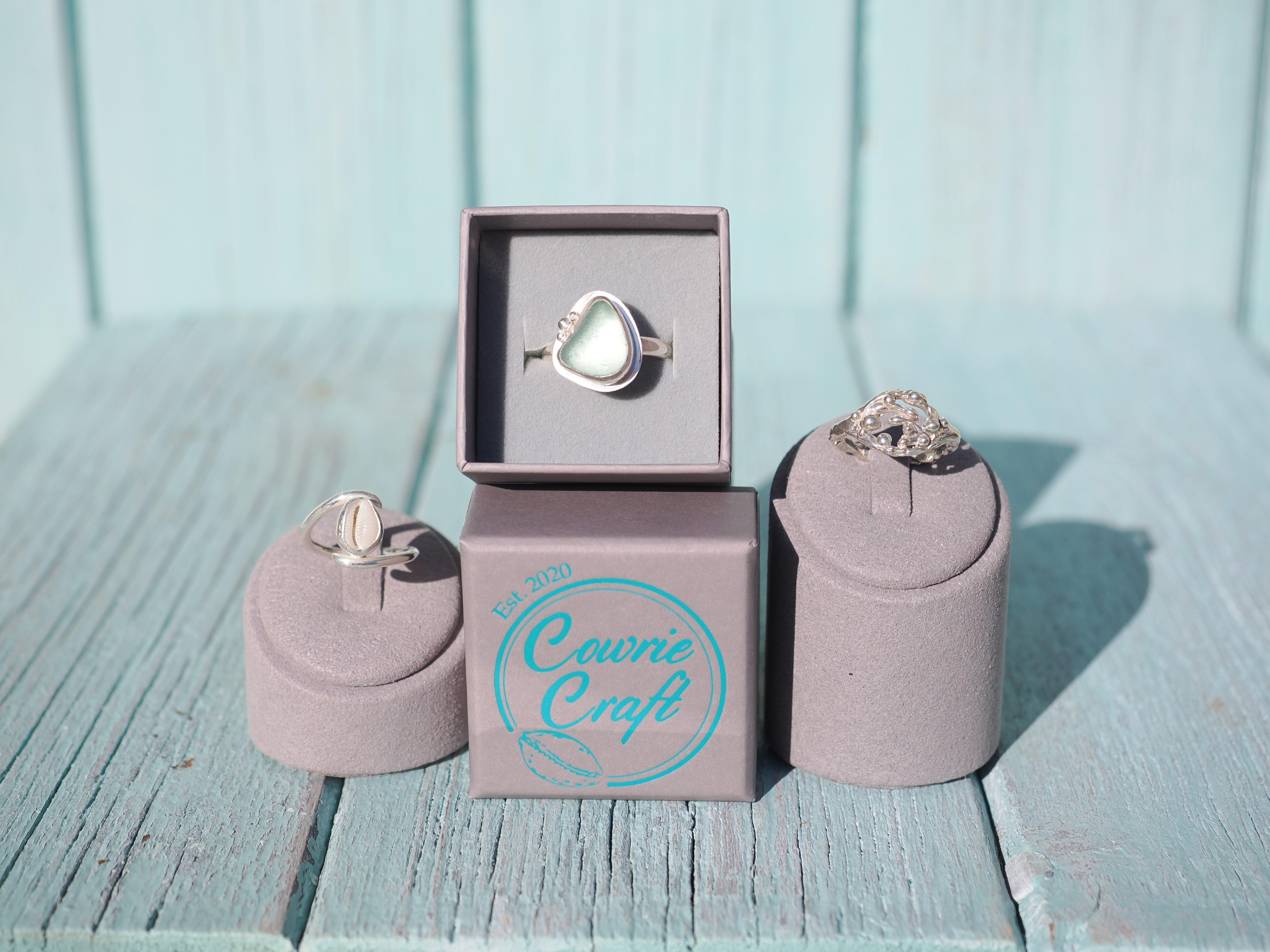 Rings resting on driftwood: sea glass, cowrie, silver Seaweed and a silver cuddy bead ring