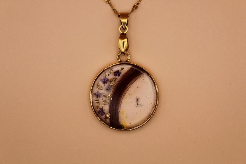 Ce pendentif rond en acier inoxydable doré met en valeur une délicate mèche de cheveux intégrée dans une résine transparente, pour un rendu léger et élégant. Quelques fleurs violettes séchées et de fines feuilles d’or viennent sublimer la création avec douceur et raffinement.

Personnalisé avec une initiale, ce bijou unique capture un souvenir précieux dans une pièce intemporelle à porter près de soi.
Un souvenir à fleur de peau