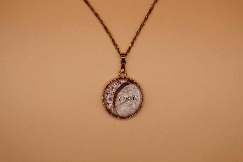 Pour cette création, ma cliente a choisi un pendentif rond en acier inoxydable doré, sur fond blanc, orné d’une mèche de cheveux, de fleurs délicates, de feuilles d’or et personnalisé avec un prénom.