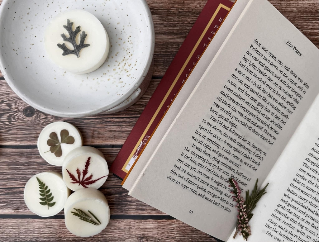 Round wax melts with botanical flowers on the top of them. They are on a wooden background with an open book on the righthand side. Wax melt collection is the title.