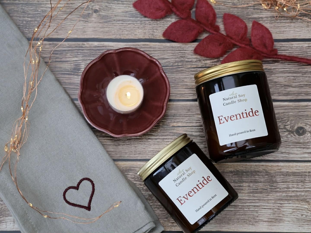Two candles in amber glass jars laying flat on a wooden background, both of the jars have natural soy candle shop on the front of them.