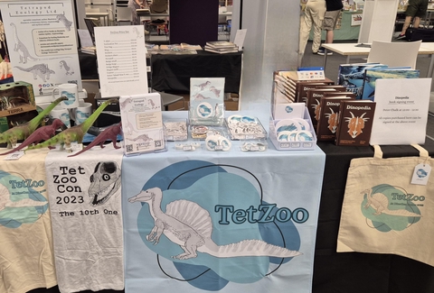 TetZoo (Tetrapod Zoology Ltd) table setup at DinoCon UK, showing the TetZoo Spinosaurus logo on T-shirts, tote bags, stickers and mugs, and a selection of books i.e Dinopedia and ANCIENT SEA REPTILES
