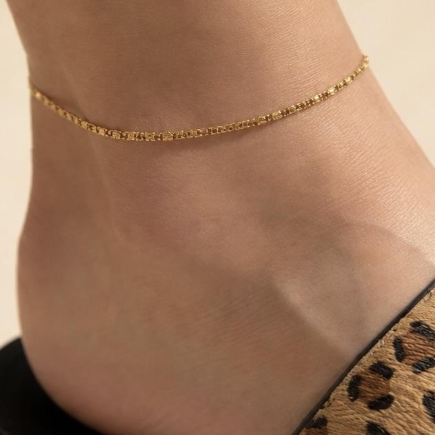 Bracelet de cheville doré en acier inoxydable porté autour de la cheville, avec une chaîne fine aux détails délicats inspirés des coquillages.