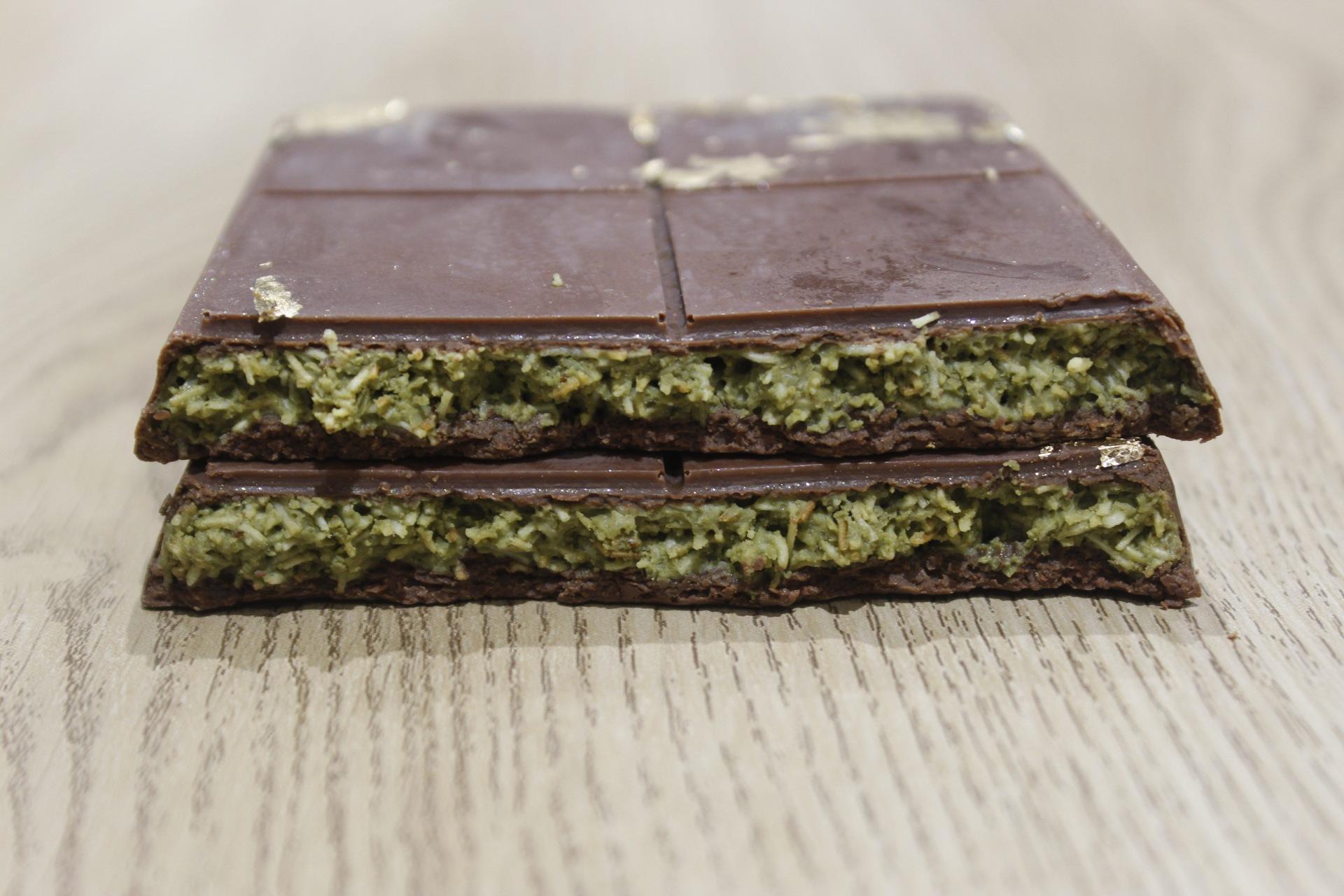 A cross-section view of two pieces of dark chocolate filled with a green, shredded filling, possibly pistachio and konafa, resting on a light wooden surface.
