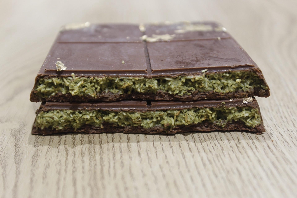 A cross-section view of two pieces of dark chocolate filled with a green, shredded filling, possibly pistachio and konafa, resting on a light wooden surface.