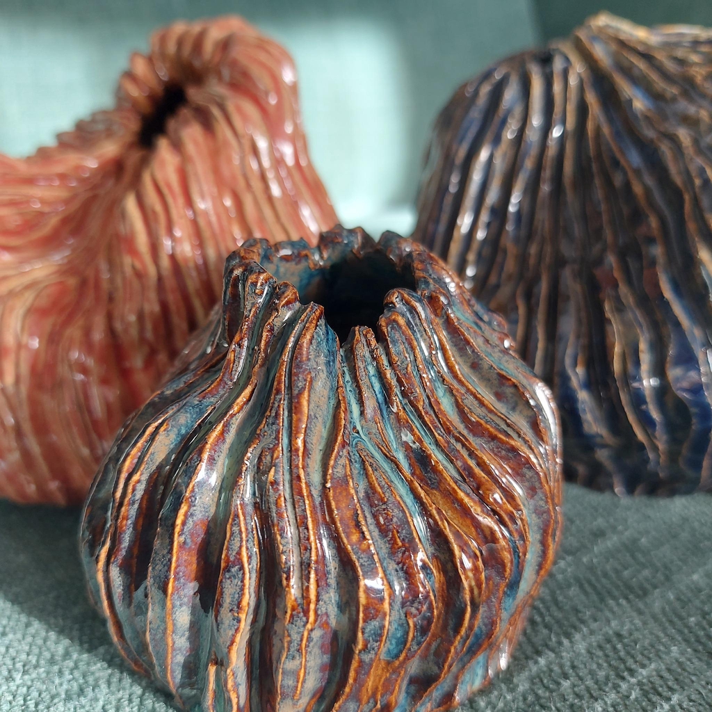a trio of small squashed vases with deep engravings. They are displayed in a triangle formation and are glazed in different colours. The back left is a coral pink, the back right is dark blue and the closest one is a mixture of browns, blues and light blue.