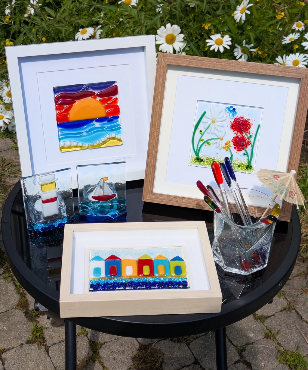 Several fused glass items displayed on a small table: framed pictures of flowers and beach huts, a tea light holder with a glass lighthouse and another with a sailboat on the sea, and a glass tumbler holding some colourful fused glass drinks stirrers.