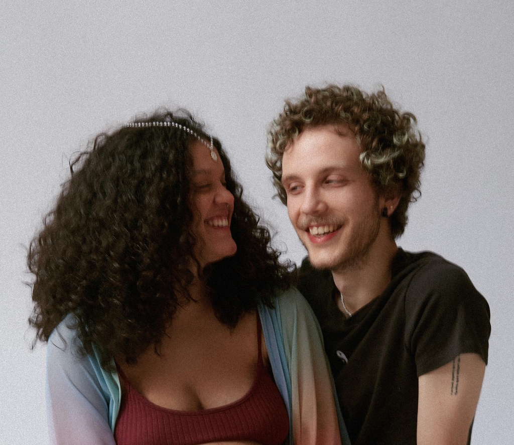 Uma foto das artistas Isabel de Toledo e Sol Soares, a Isabel é a primeira a aparecer na foto (estando no lado esquerdo, na frente). Ela tem o cabelo cacheado comprimido, usa um acessório de cabelo branco, um top bordô e um kimono arco íris. Já a Sol que, se encontra atrás da Isabel, tem o cabelo curto cacheado e loiro. Está usando uma camiseta preta com uma gargantilha de pérolas.