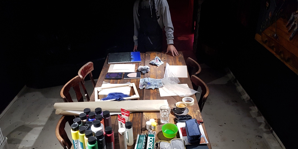 Photo of a table with art materials and the artist standing.