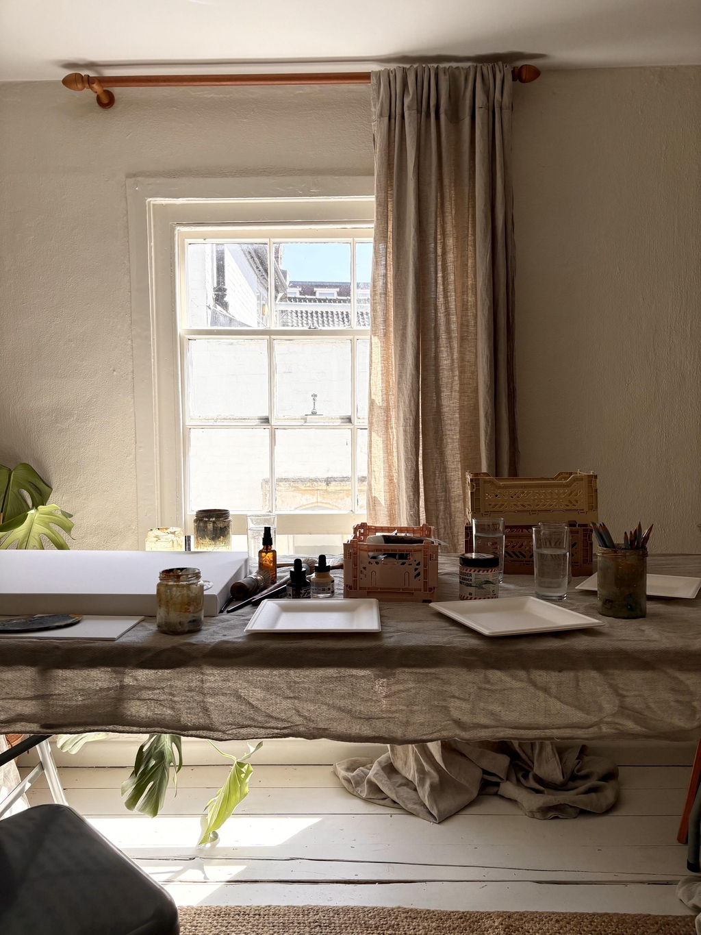 A sunlit workspace with art supplies, jars, and white plates on a linen-covered table in front of a window with beige curtains.