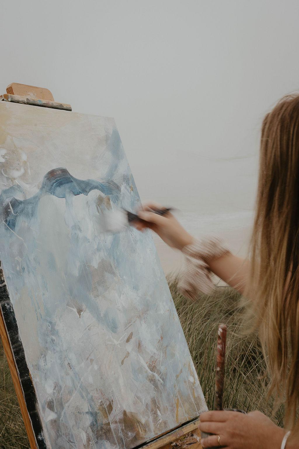 An unfinished painting on an easel at a beach. The artist's hand is holding a thick brush making strokes across the canvas. 