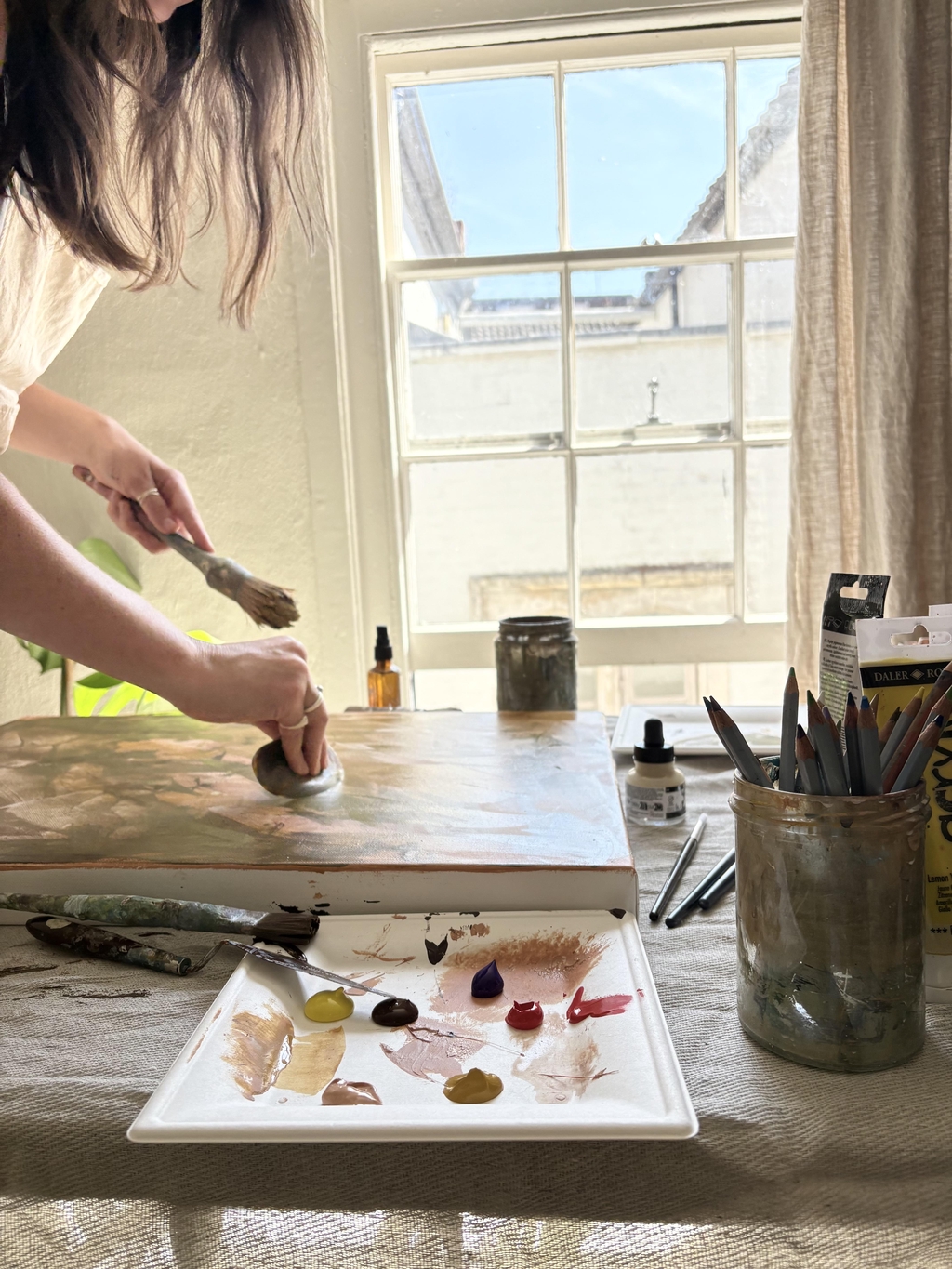 A woman working on a stretched canvas on a table. Paint, pencils and brushes are next to it. An open window in the background.