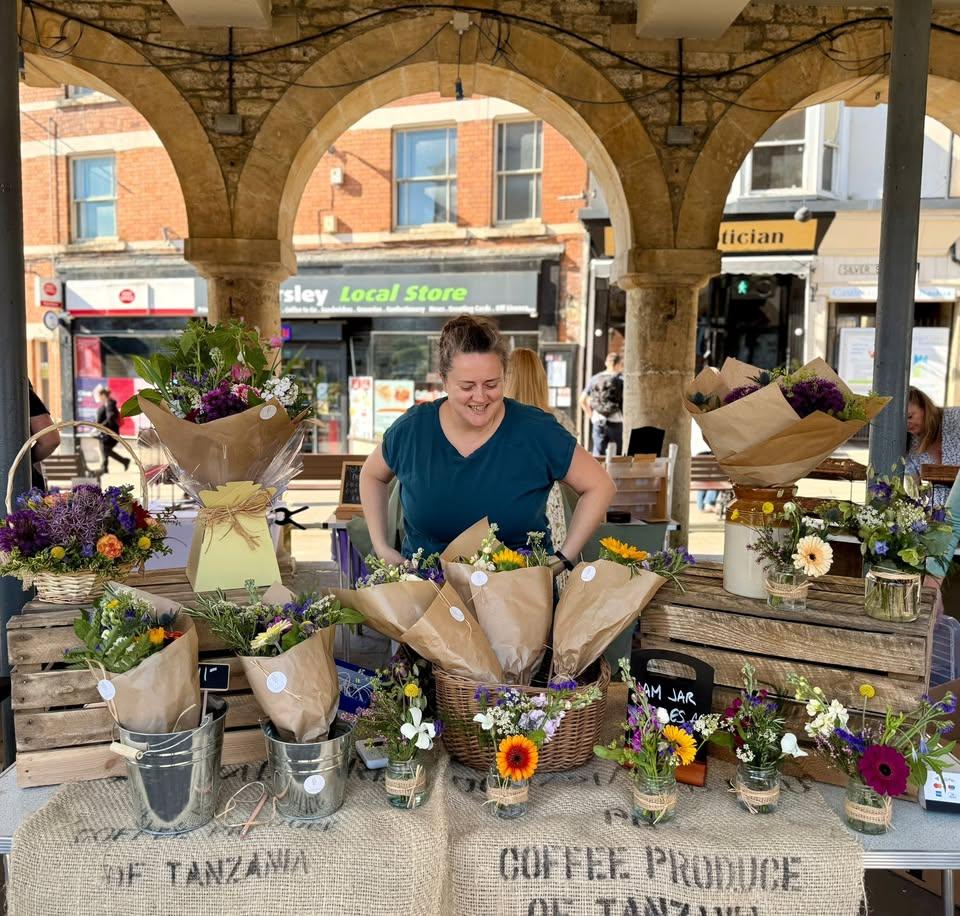 Sophie standing behind her stall at Dursley market. There are many brightly coloured bouquets and jam jar posies on the stall.