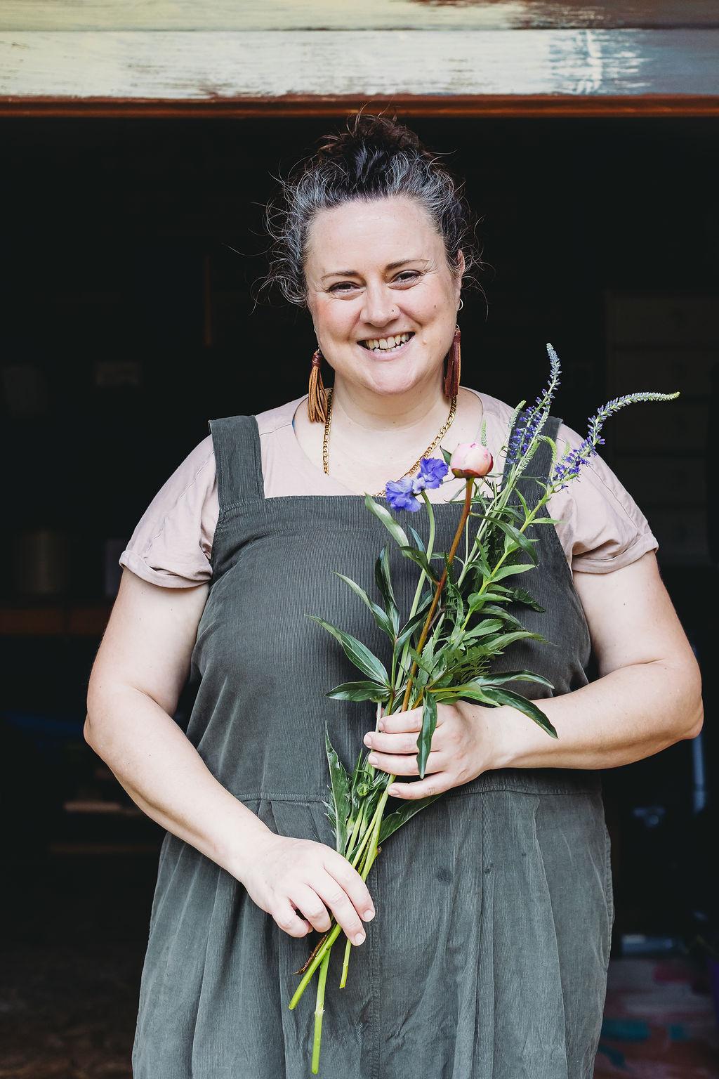 Sophia, a middle aged white woman, is grinning at the camera. In the foreground she holds a bunch of pink and purple flowers.