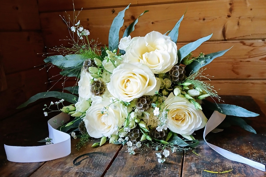 A bridal bouquet of ivory roses surrounded by smaller roses, lisianthus and lots of textural foliage, grasses and small flowers. It is finished with white ribbon.
