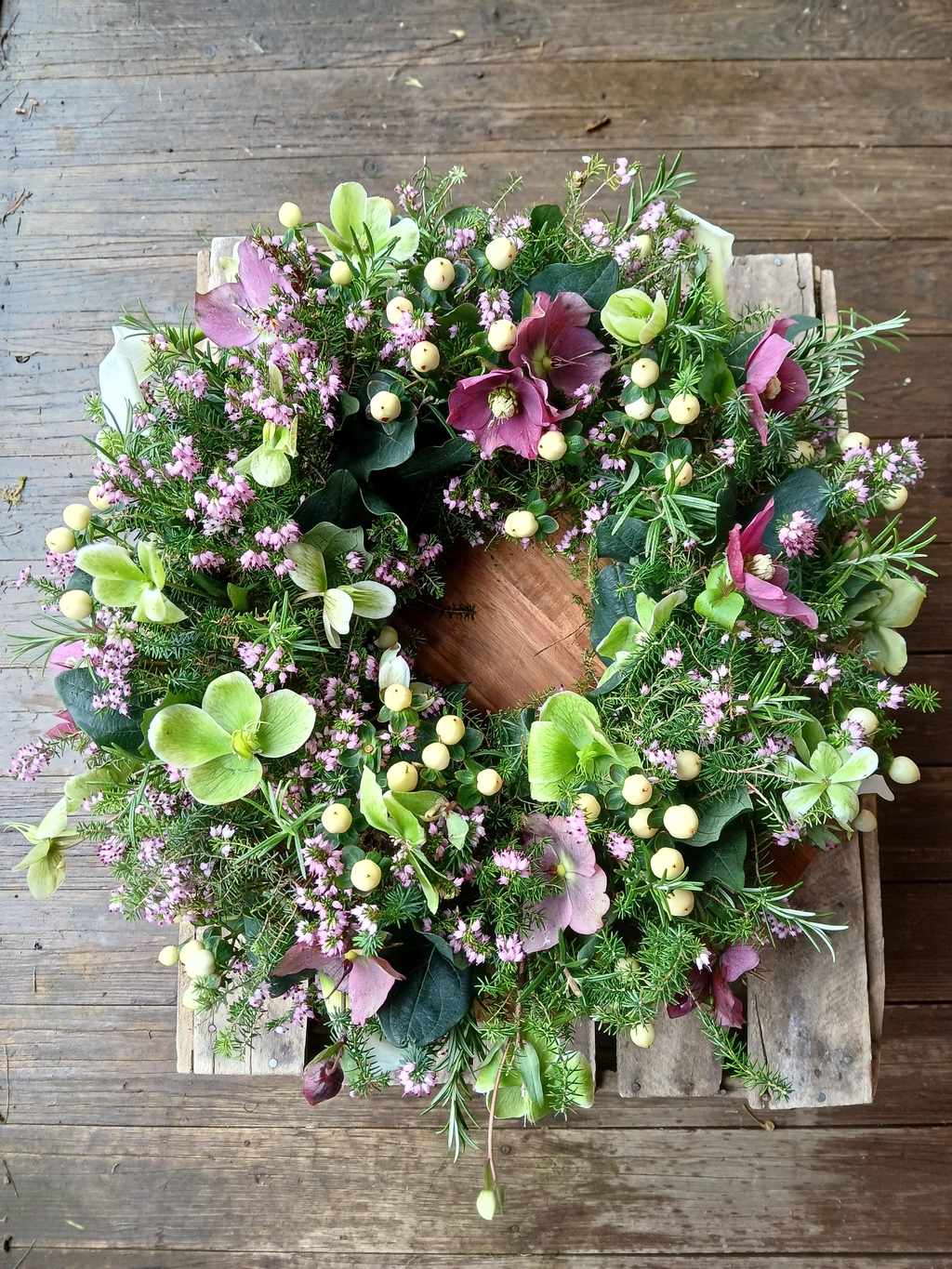 A sustainable funeral wreath featuring purple and green hellebores, white hypericum and pink heather, with rosemary and heather foliage.